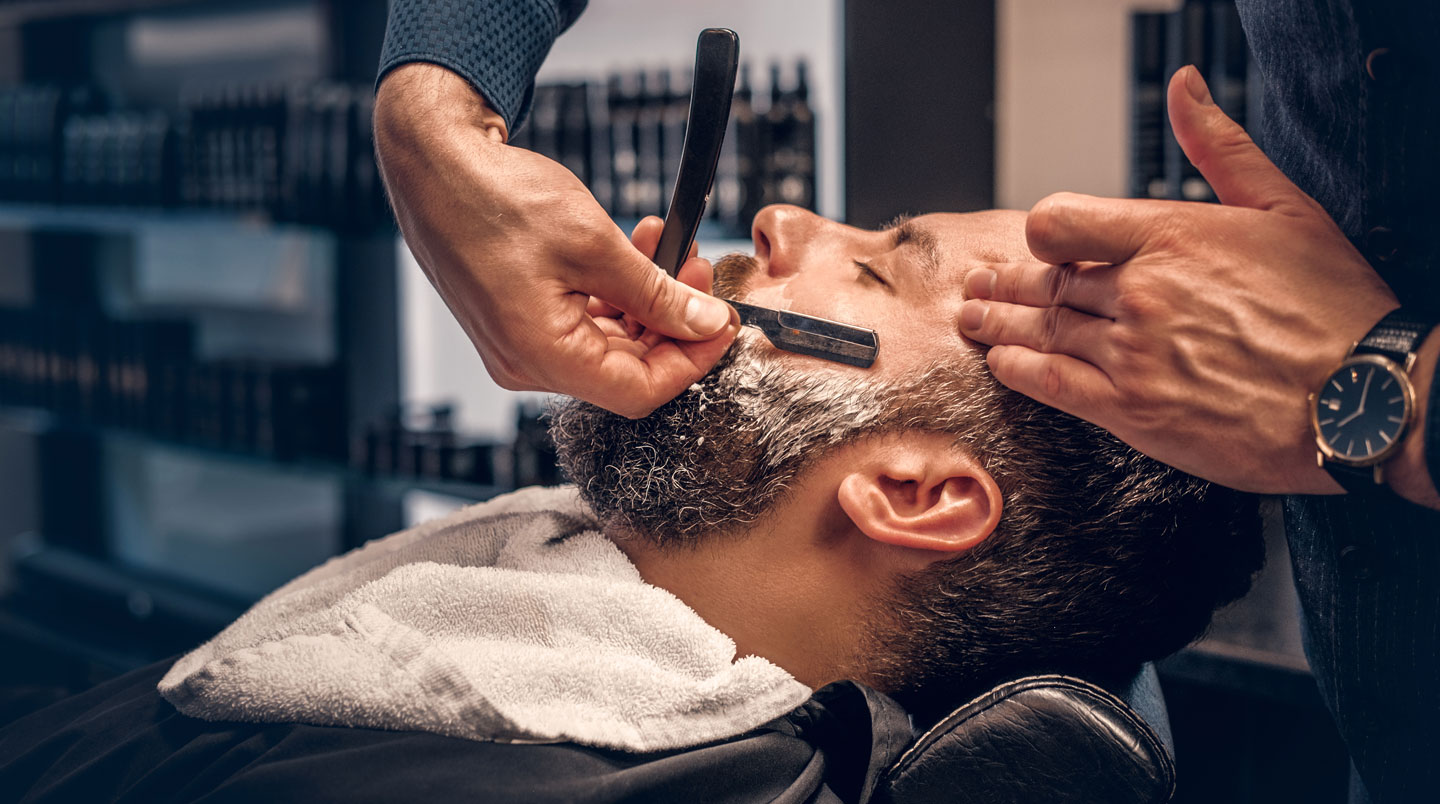 Gros plan de profil sur une joue masculine pendant le rasage, montrant un rasoir coupe-choux positionné à angle faible contre la peau, avec zone partiellement rasée visible et main tenant correctement le rasoir